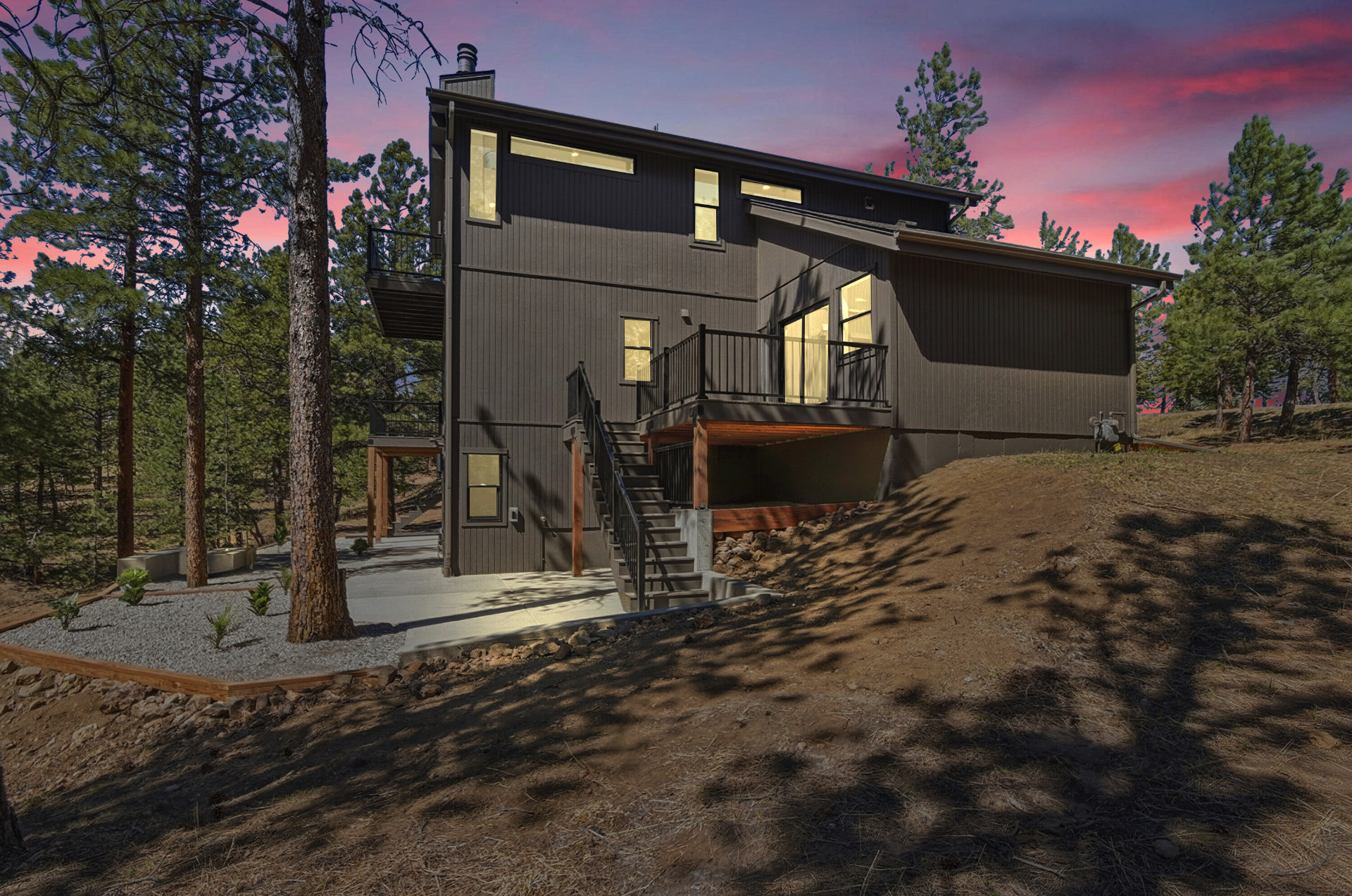 Modern mountain home at dusk with glowing windows, a wood deck, and pine trees